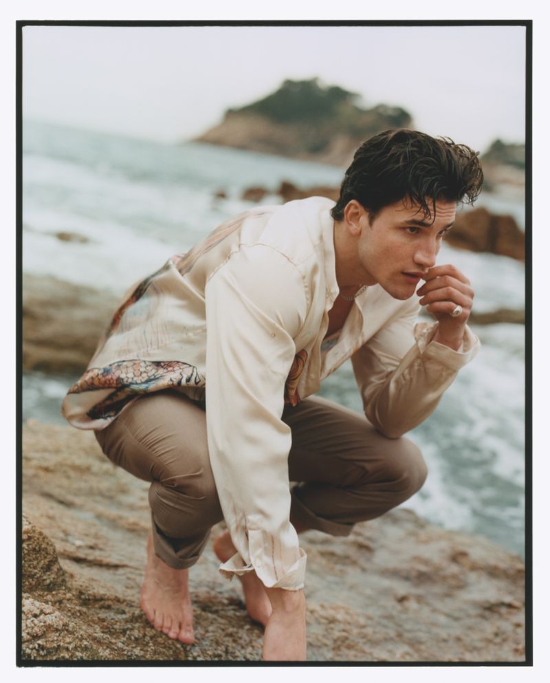 Man in an open shirt squatting on rocks by the ocean, with island in the background on an overcast day, creating a contemplative mood.
