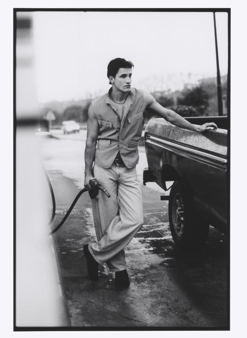 Monochrome portrait of a man in a vest leaning against a truck at a gas station, holding a fuel pump nozzle in his hand.