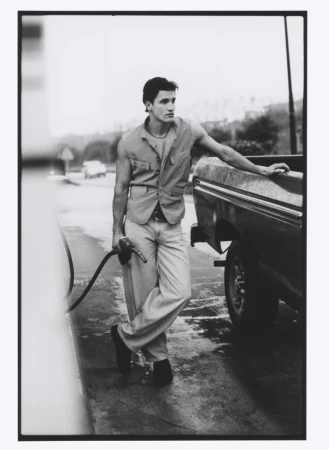 Monochrome portrait of a man in a vest leaning against a truck at a gas station, holding a fuel pump nozzle in his hand.