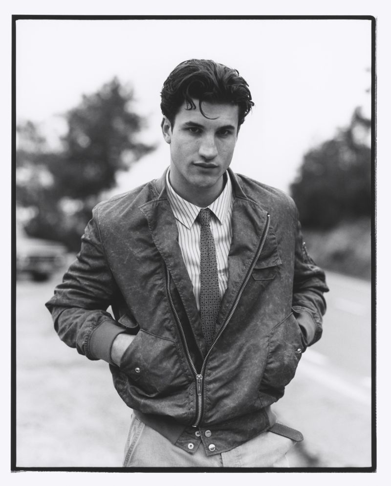 Monochrome portrait of a young adult male wearing a jacket, striped shirt and tie standing on a roadside with trees in the background.