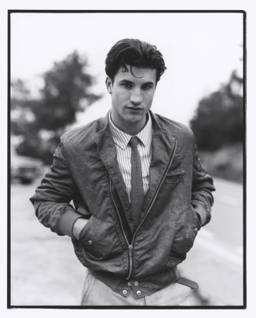 Monochrome portrait of a young adult male wearing a jacket, striped shirt and tie standing on a roadside with trees in the background.