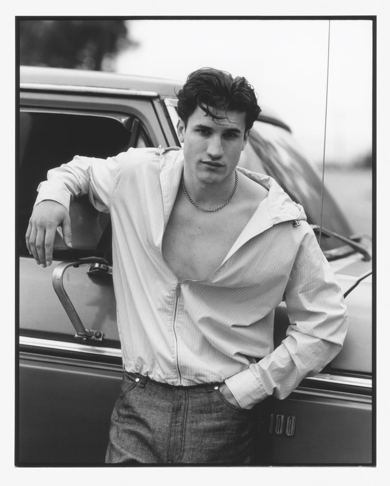 Monochrome portrait of a young adult male leaning against a vehicle, wearing an unbuttoned shirt and jeans in an outdoor setting.
