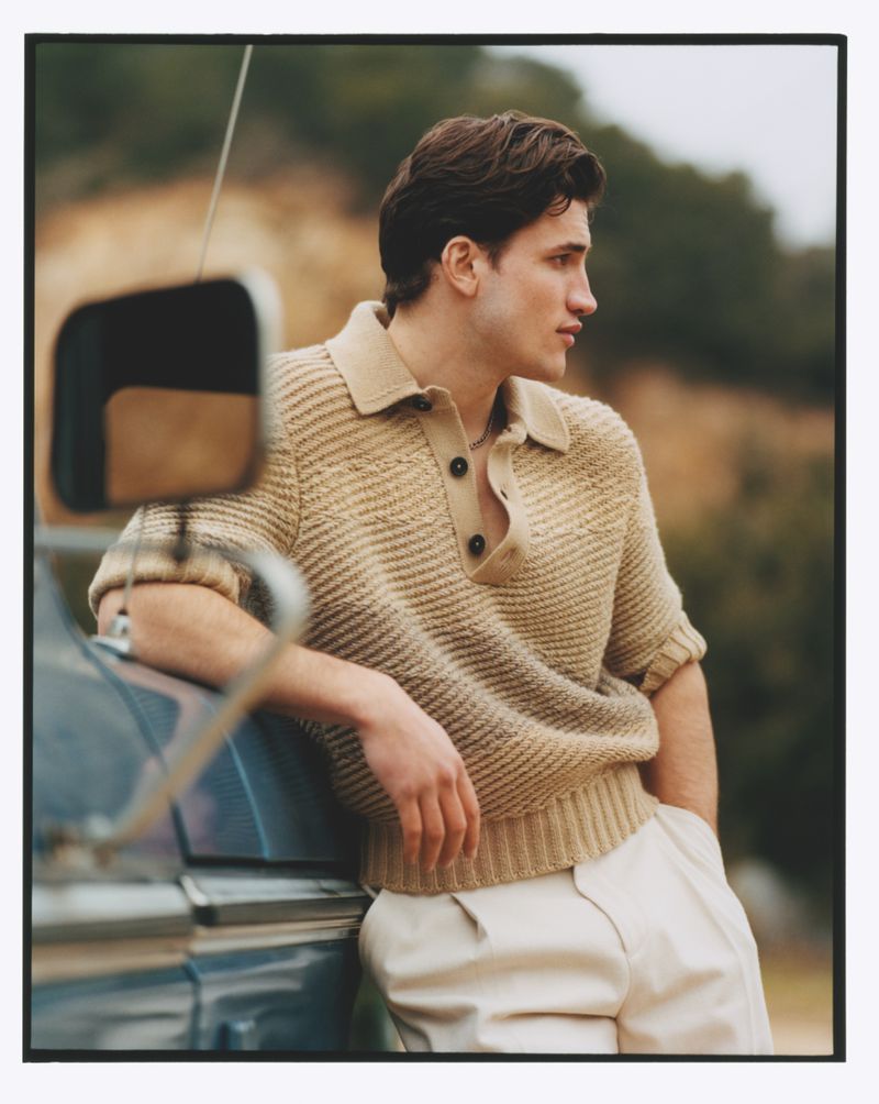 Young man in a tan short-sleeve knit shirt and white pants leans against a blue car with a natural outdoor background. Portrait orientation.