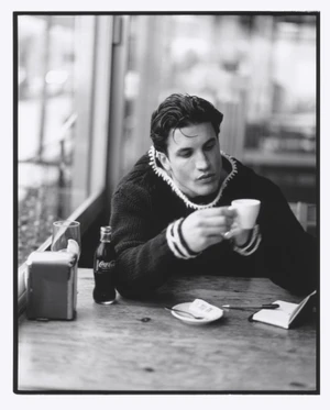 Max Knott seated at a wooden café table holding a coffee cup, photographed by James Harvey-Kelly for Numero Homme.