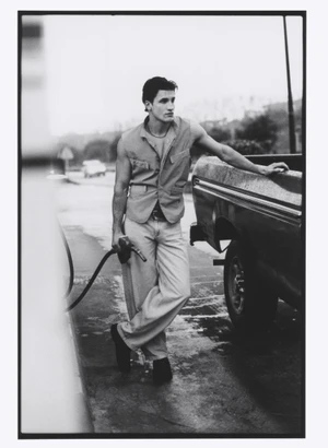 Black and white editorial image of a male model leaning against a pickup truck holding a gas pump nozzle at a service station