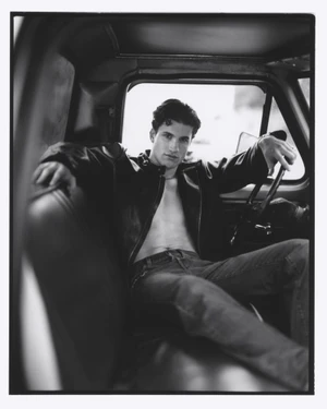 Black and white portrait of a male model seated inside a truck cab wearing a dark leather jacket and jeans.