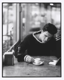 Monochrome portrait of a young man seated at a wooden table in a cafe, resting his head on his hand next to a coffee cup and glass bottle.