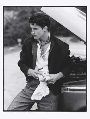 Monochrome portrait of a young man wearing a patterned neckerchief leaning against an open car hood outdoors.
