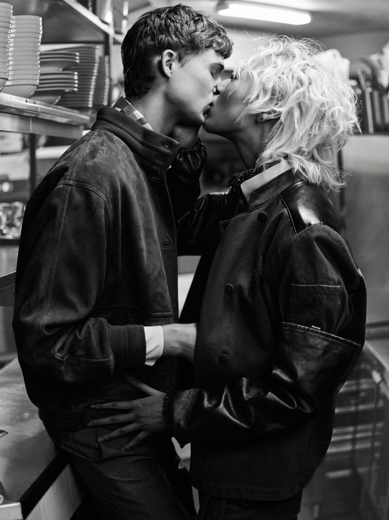 A monochrome portrait captures a couple kissing in what appears to be a kitchen, both in jackets with soft lighting and a shallow depth of field.