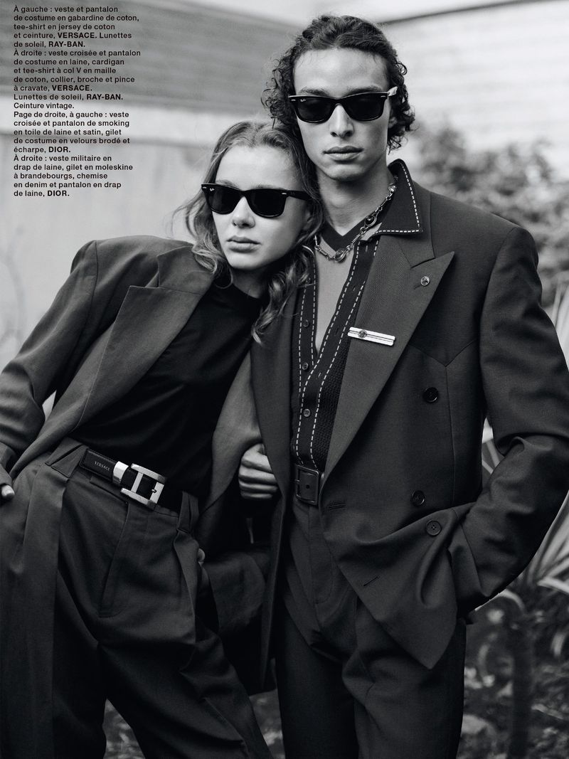 Two fashion models in sunglasses pose in matching suits. The female model has her arm around the male model in black and white.