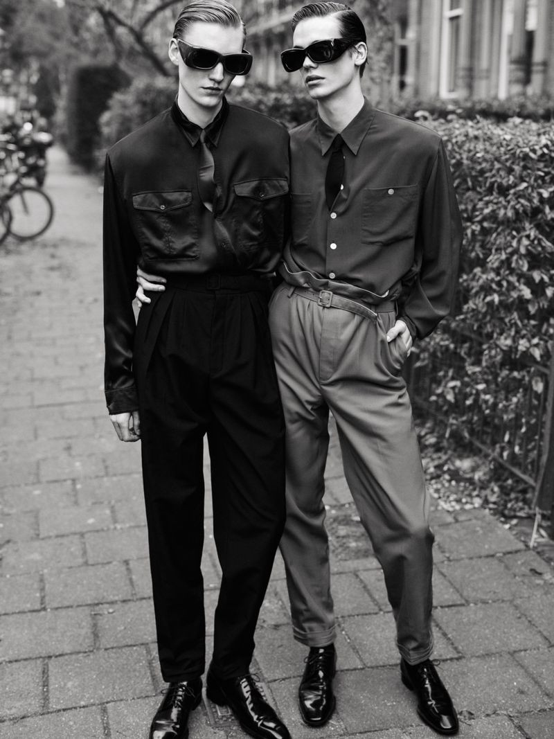 Two young men in vintage-style clothing pose on a brick walkway, wearing sunglasses and formal attire. A bicycle is visible in the background.