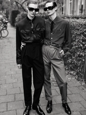 Two young men in vintage-style clothing pose on a brick walkway, wearing sunglasses and formal attire. A bicycle is visible in the background.