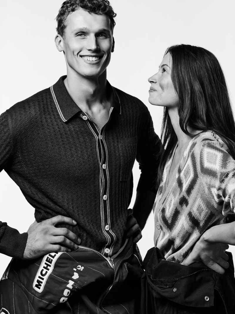 Black and white portrait of a smiling man and woman in racing gear against a white background, hands on hips, looking at each other.
