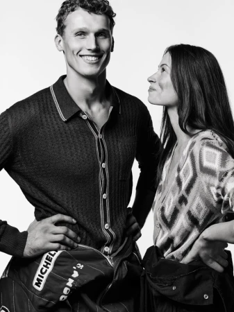 Black and white portrait of a smiling man and woman in racing gear against a white background, hands on hips, looking at each other.