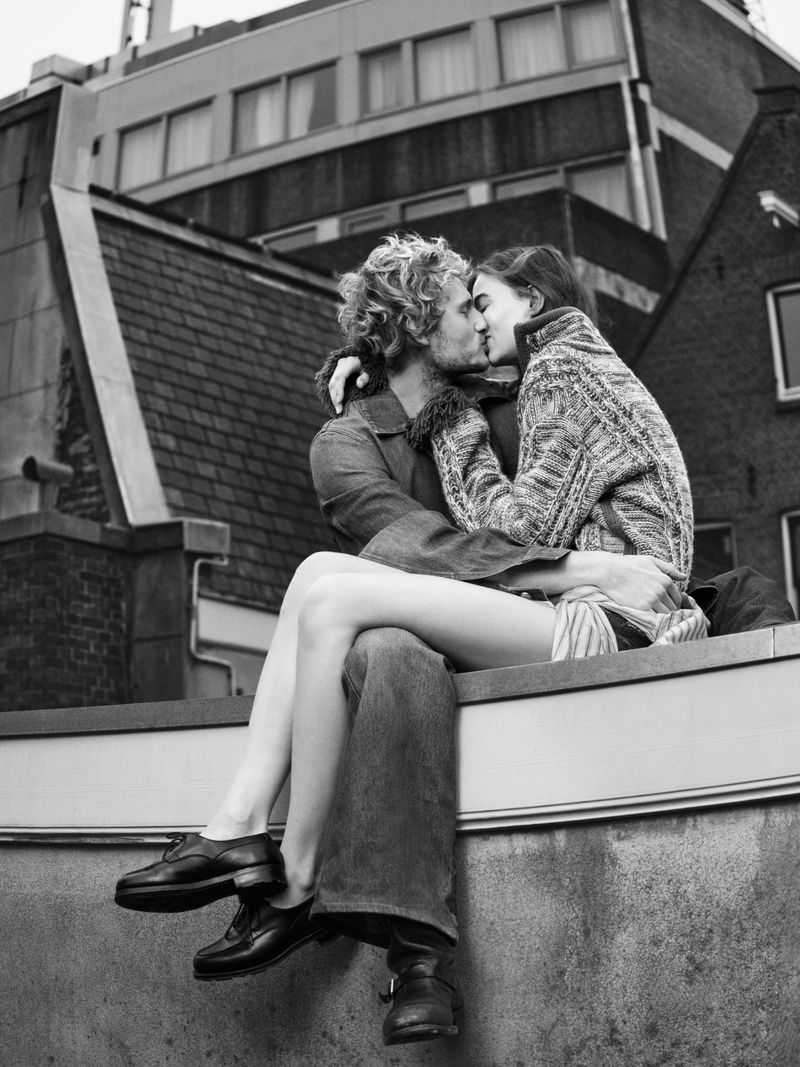 Black and white shot of couple kissing on a rooftop, embracing in sunlight, with a cityscape and building in the background.