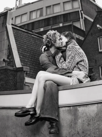 Black and white shot of couple kissing on a rooftop, embracing in sunlight, with a cityscape and building in the background.