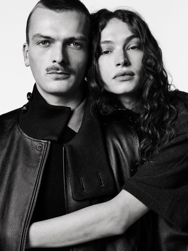 Black-and-white portrait of a couple in a close embrace. The man has a mustache, and the woman has long, curly hair and a serious facial expression.