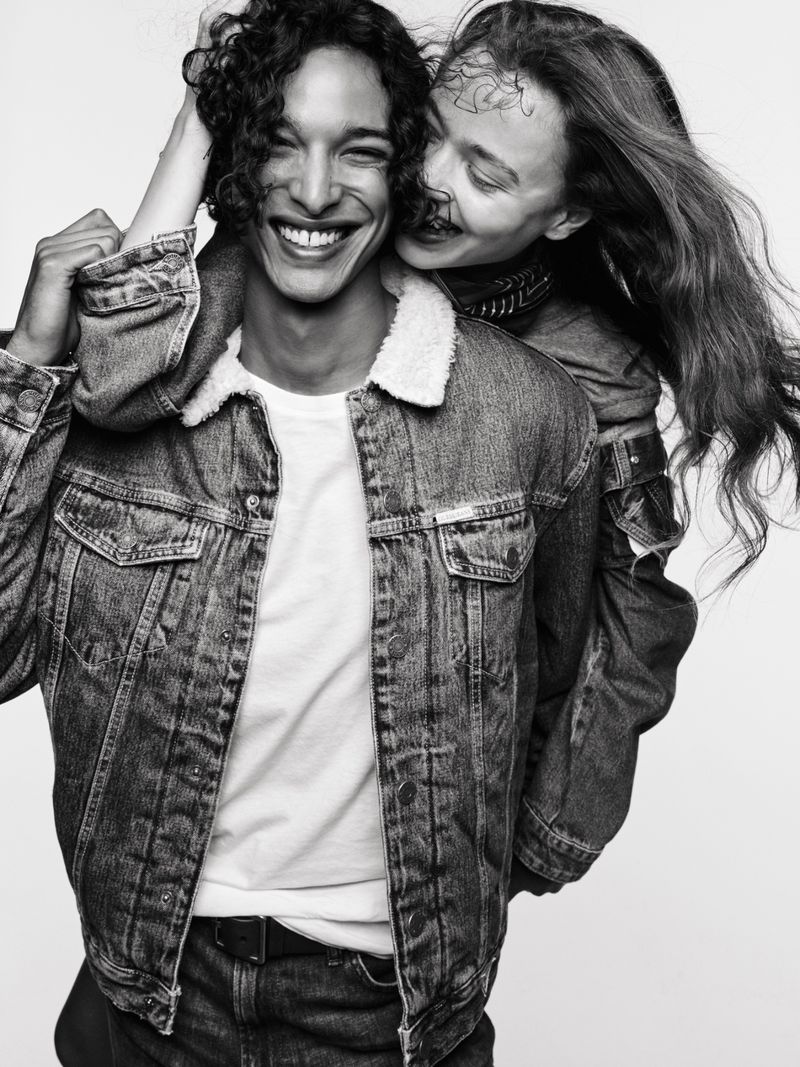 Black and white studio shot of an attractive man and woman. The couple wears denim jackets and smiles while posing against a plain background.