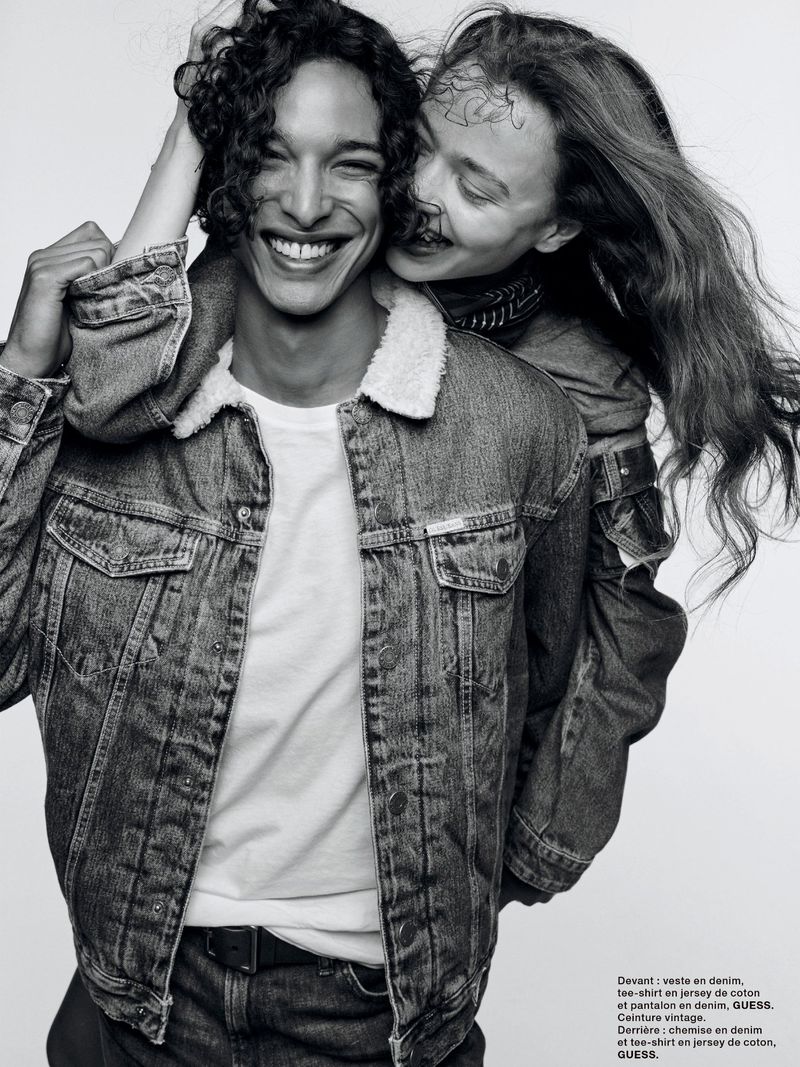 A smiling man in a denim jacket and white t-shirt is embraced from behind by a woman in a similar jacket, set against a white background.