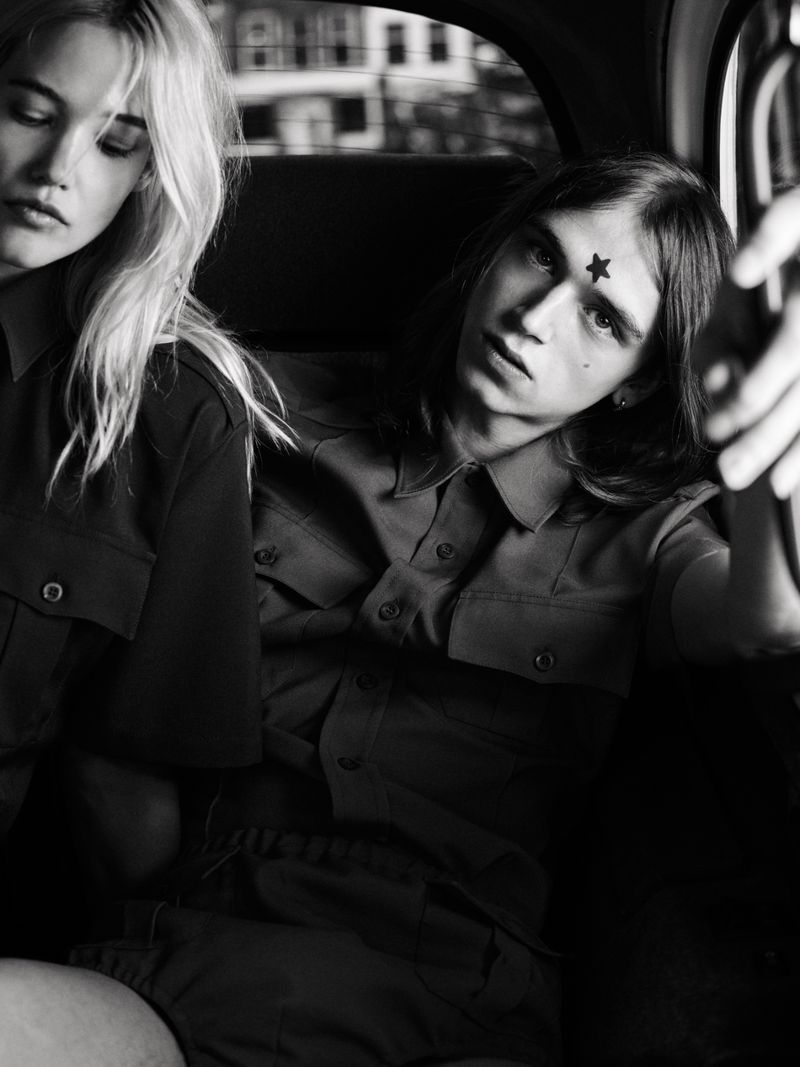 Black and white portrait of a blond woman and a man with a star on his forehead in a car, both wearing button-down shirts.
