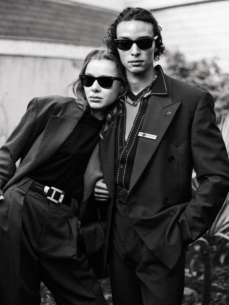 Monochrome portrait of a stylish couple, embracing in blazers and sunglasses, set against a neutral urban outdoor background.