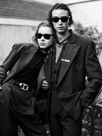 Monochrome portrait of a stylish couple, embracing in blazers and sunglasses, set against a neutral urban outdoor background.