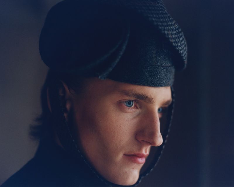 Close-up portrait of a young adult male with blue eyes wearing a dark-colored houndstooth cap and dark shirt against a dark background.