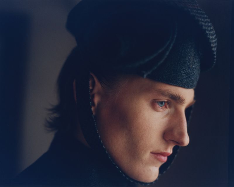Portrait of a fair-skinned person with blue eyes and dark hair, wearing a dark hat and a dark coat in a dimly lit indoor environment.