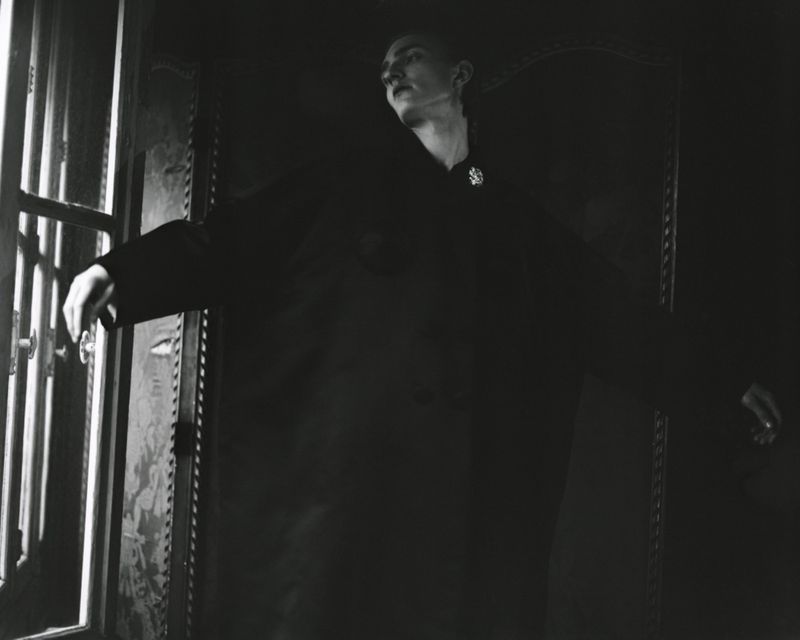 Monochrome image of a slender person in dark clothing by a window, face turned to the side, hands extended, creating a dramatic, artful portrait.