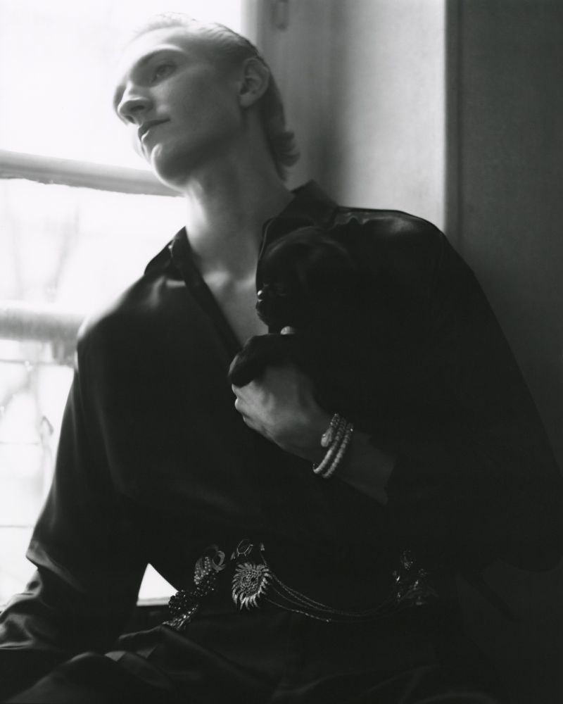 Black and white portrait of a slender adult male with bracelets and an ornate belt sitting in front of a window.