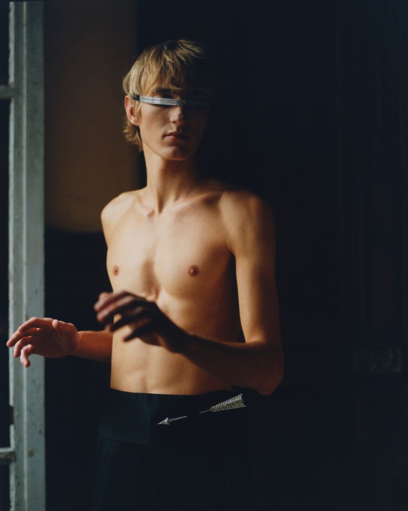 Young man with blond hair and futuristic visor stands shirtless in front of a window in dramatic lighting, showcasing dark athletic pants.