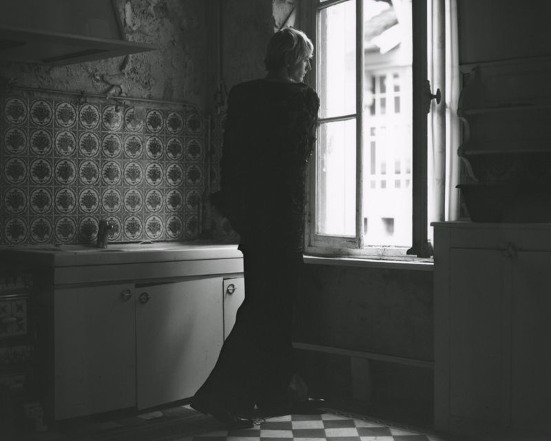 Monochrome interior shot of a woman looking out a window in a rustic kitchen with patterned tiles and checkered floor in diffused light.