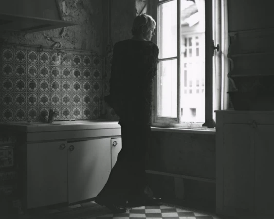 Monochrome interior shot of a woman looking out a window in a rustic kitchen with patterned tiles and checkered floor in diffused light.