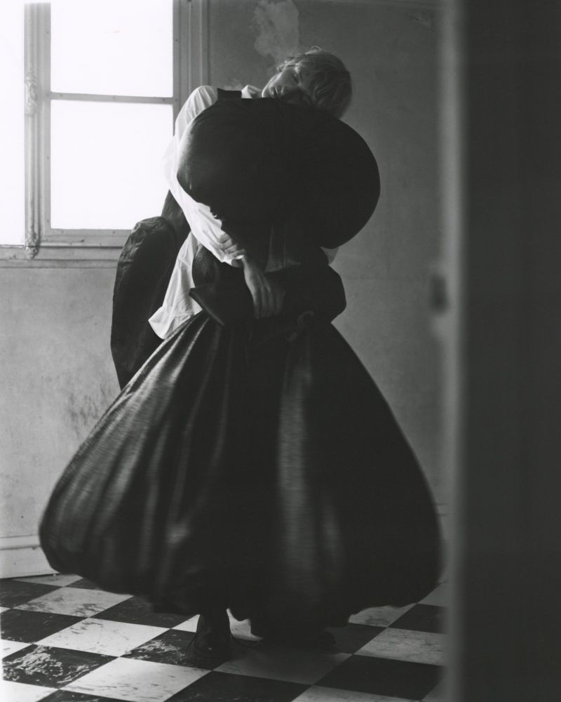 Monochrome photograph of a person wearing a white shirt and a large black skirt, indoors, standing on a checkered floor near a window.