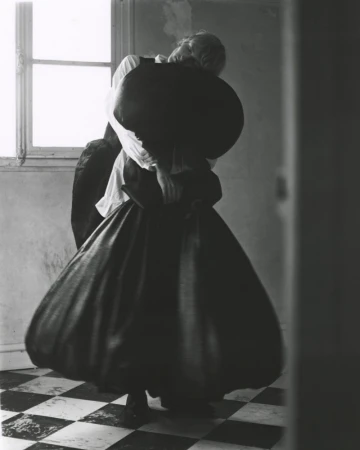 Monochrome photograph of a person wearing a white shirt and a large black skirt, indoors, standing on a checkered floor near a window.