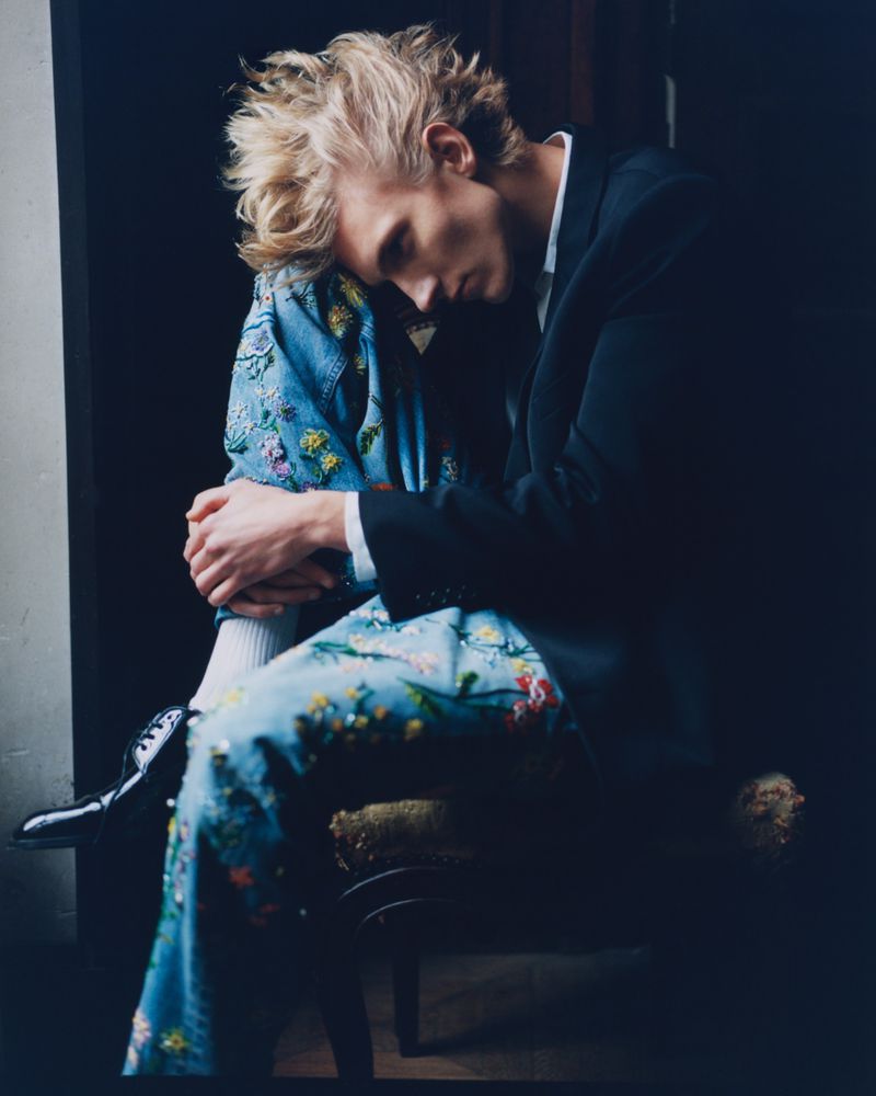 Blond male model wearing a navy blazer and patterned pants sitting in dark room, head bowed, hugging his knees in a studio shot.