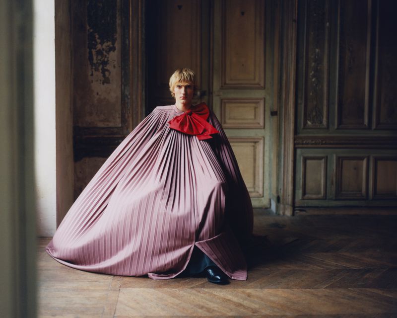 A person with blonde hair kneeling on a wooden floor wearing a large, light-purple, pleated garment with a red bow indoors.