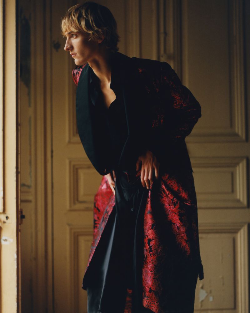 Side-view shot of a young adult man with blond hair wearing a red and black patterned jacket in front of cream-colored doors.