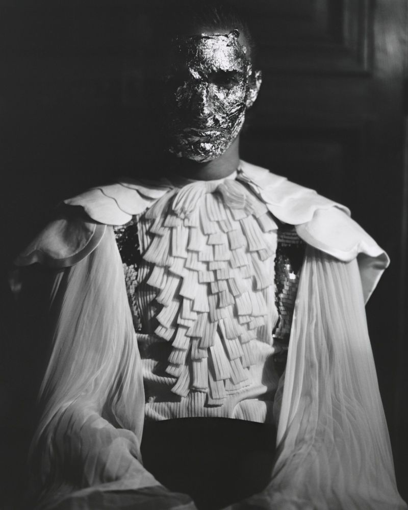Person with a metallic face covering poses indoors wearing a white ruffled collar and sheer shoulder coverings in a high-contrast black and white portrait.