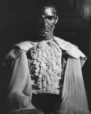 Person with a metallic face covering poses indoors wearing a white ruffled collar and sheer shoulder coverings in a high-contrast black and white portrait.