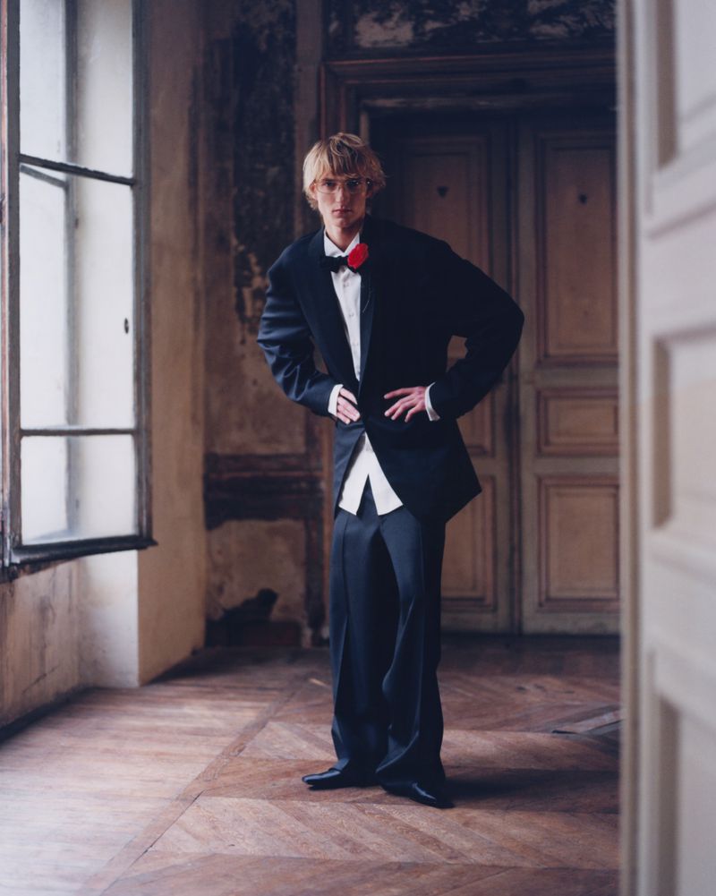 Man in black tuxedo with red bow tie poses indoors with hands on hips, standing on parquet floor near large window and paneled doors.