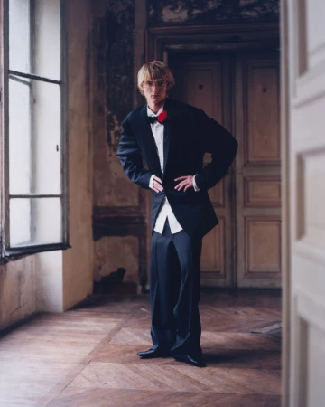 Man in black tuxedo with red bow tie poses indoors with hands on hips, standing on parquet floor near large window and paneled doors.