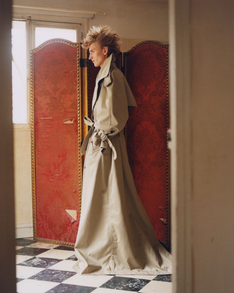 Profile view of an adult wearing a long trench coat with a red folding screen backdrop and a black and white checkered floor.