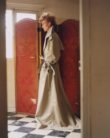 Profile view of an adult wearing a long trench coat with a red folding screen backdrop and a black and white checkered floor.