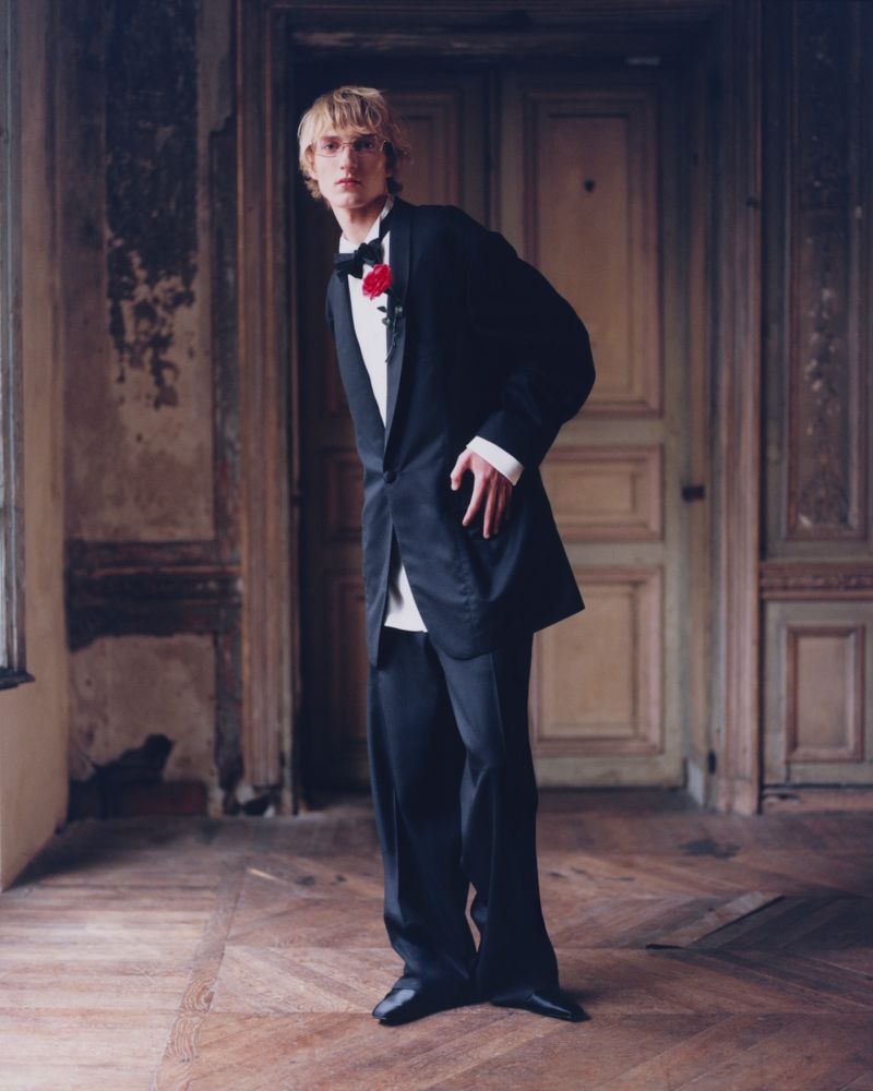 A blonde-haired man in a black tuxedo stands in an old room with ornate walls and herringbone wood flooring. Fashion editorial photography.