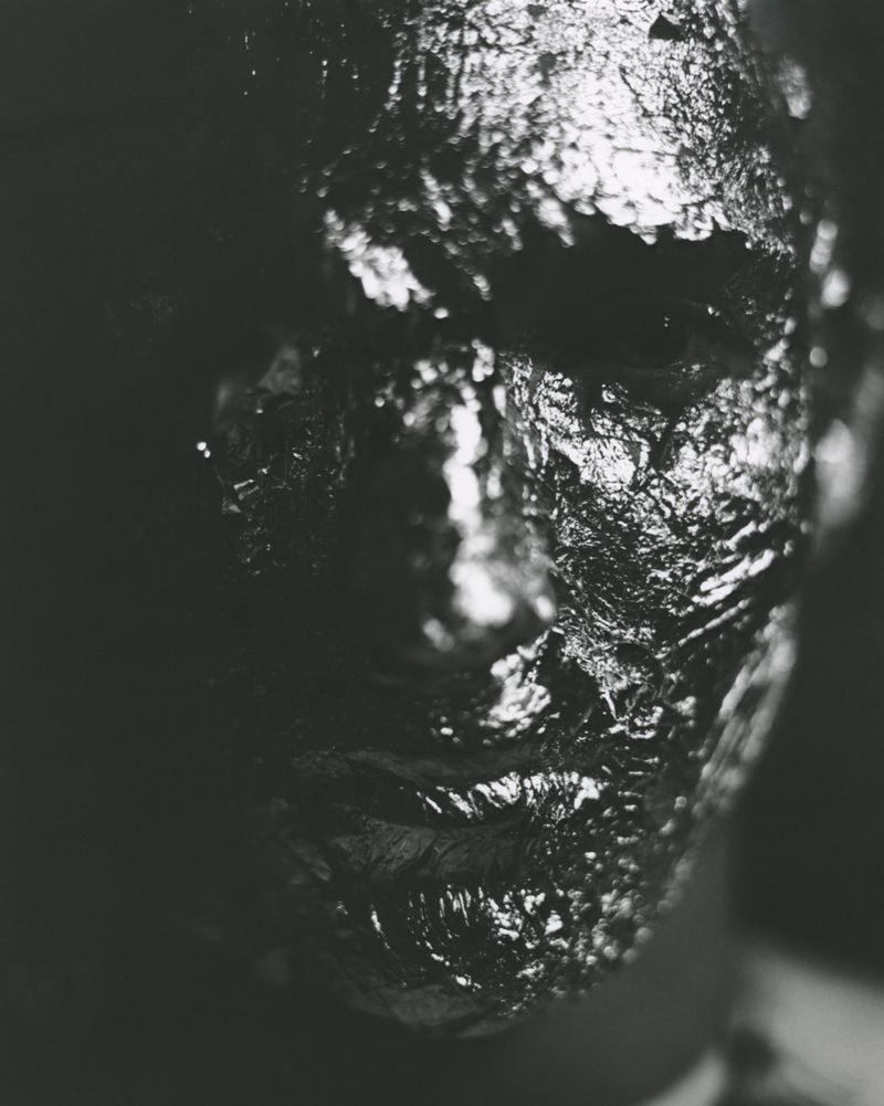 Monochrome portrait of a human face completely covered in shiny, reflective material in an indoor environment with dark, moody lighting.