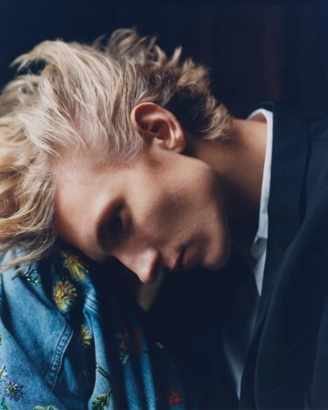 Portrait of a blonde male model resting his head against an embroidered denim jacket and wearing a dark blazer.