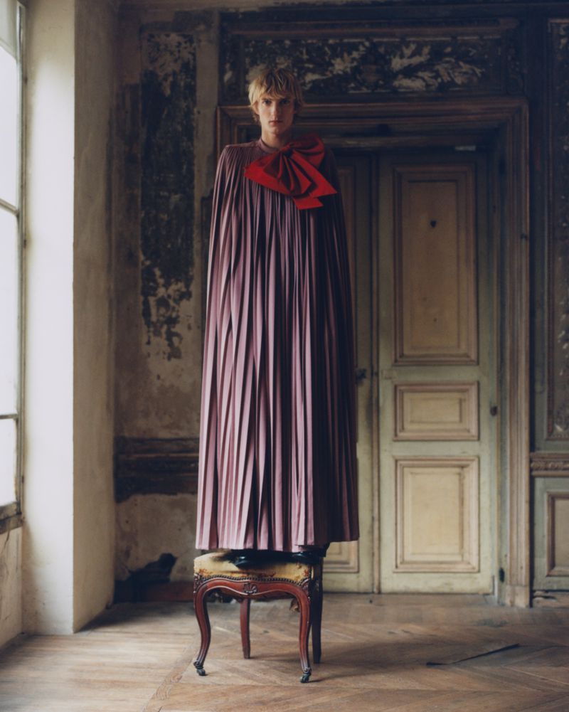 Fashion shot featuring a model wearing a long, pleated dress with a red bow, standing on a stool in a worn, vintage-style interior.