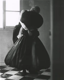 Model in voluminous dark gown holding a large black sculptural form near a window with checkered flooring