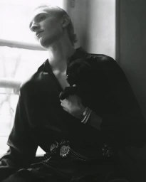 Monochrome portrait of a male model wearing a dark shirt and ornate belt while holding a puppy near a bright window